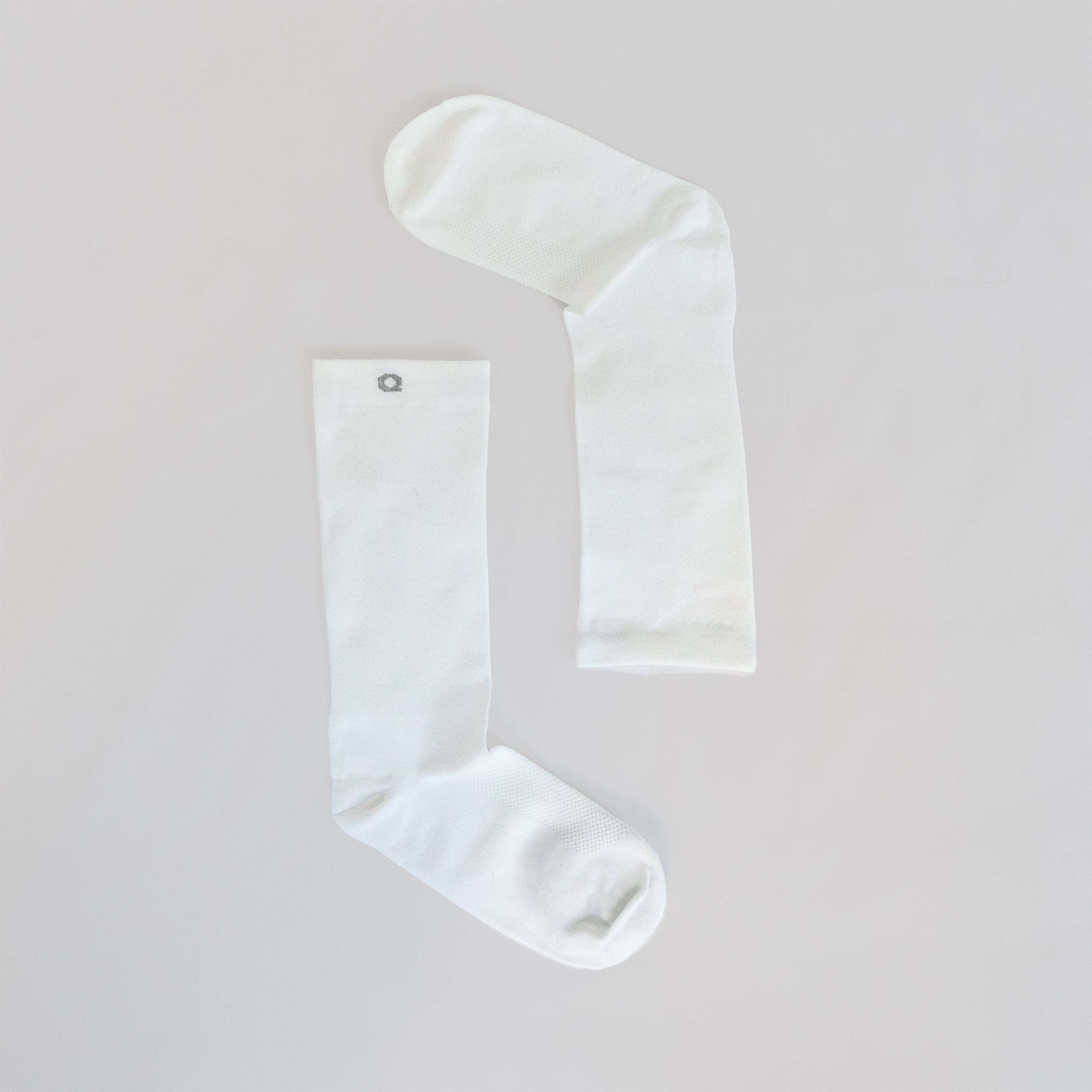 Two plain white socks laid flat on a light surface, positioned to form an angled shape resembling an upside-down letter L. One sock has a small gray circular emblem near the top.